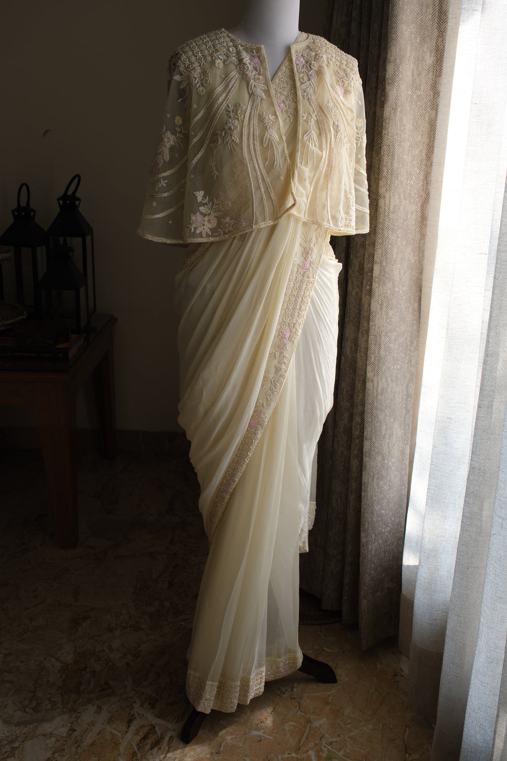 A cream-colored traditional sari with floral embroidery and a matching embroidered blouse displayed on a mannequin in a dimly lit room next to a window.