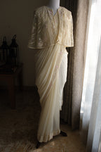 A cream-colored traditional sari with floral embroidery and a matching embroidered blouse displayed on a mannequin in a dimly lit room next to a window.