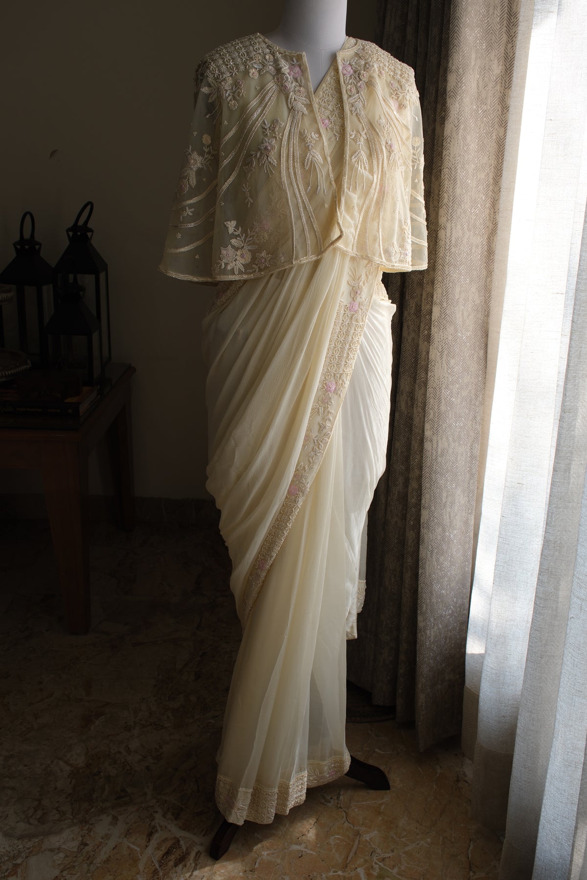 A cream-colored traditional sari with floral embroidery and a matching embroidered blouse displayed on a mannequin in a dimly lit room next to a window.