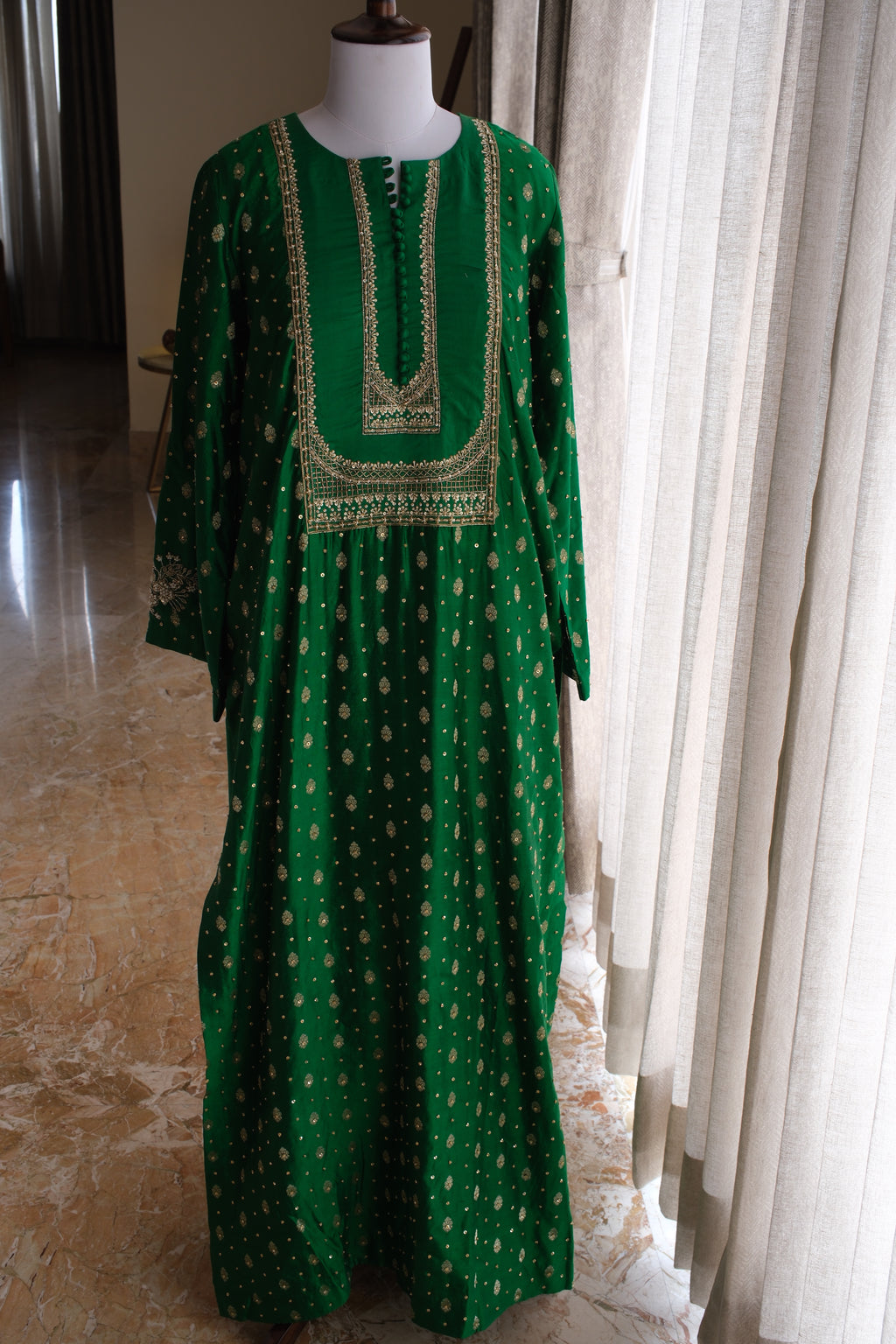 A green long-sleeved tunic dress with gold geometric patterns and a gold embroidered neckline, displayed on a mannequin in an indoor setting with curtains and a tiled floor.