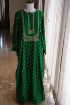 A green long-sleeved tunic dress with gold geometric patterns and a gold embroidered neckline, displayed on a mannequin in an indoor setting with curtains and a tiled floor.