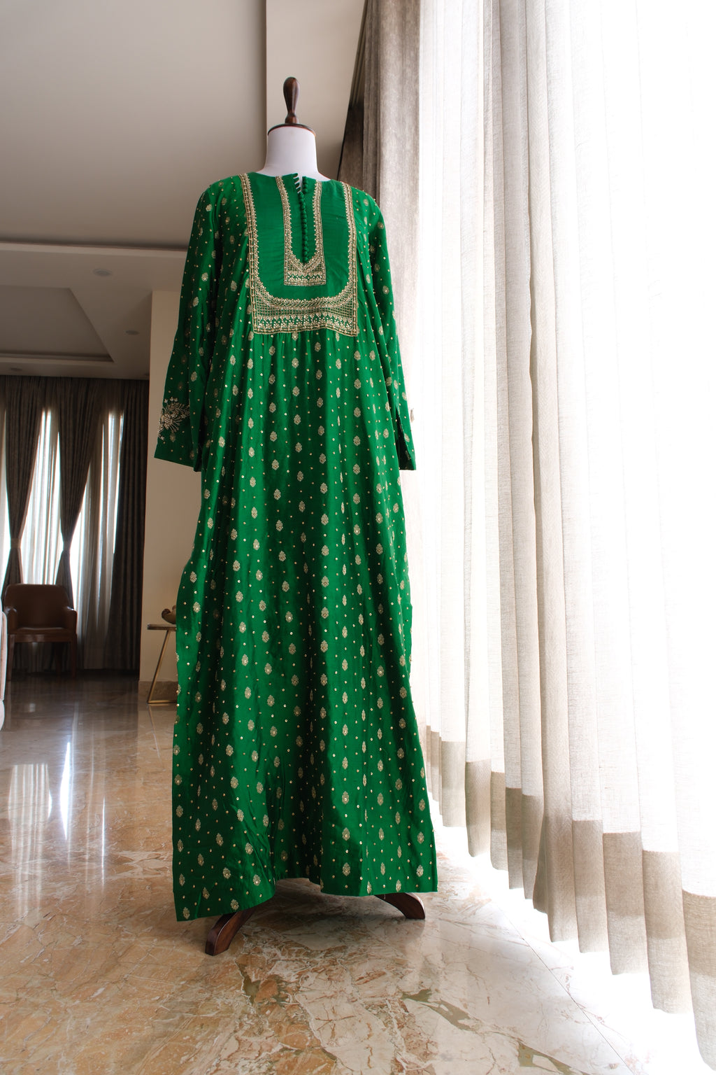 A long green kaftan dress with gold patterns and embroidery, displayed on a mannequin in a bright room with curtains and marble flooring.