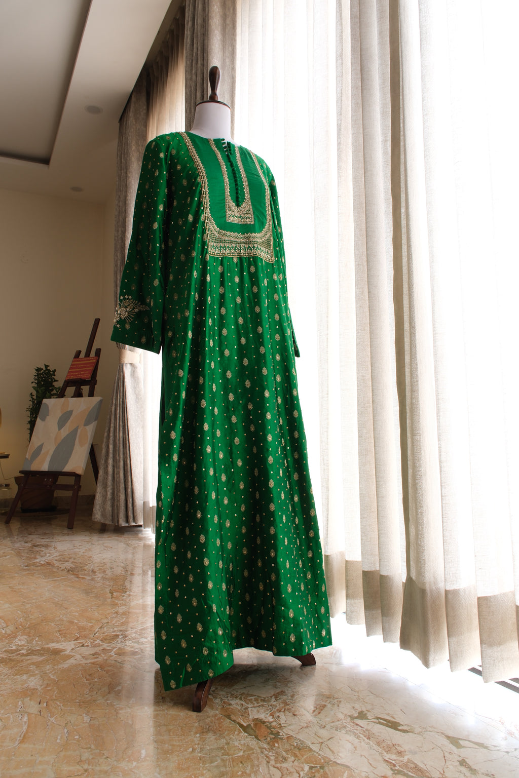 A long green kaftan dress with gold geometric patterns and a gold embroidered neckline, displayed on a mannequin in a bright room with white curtains.