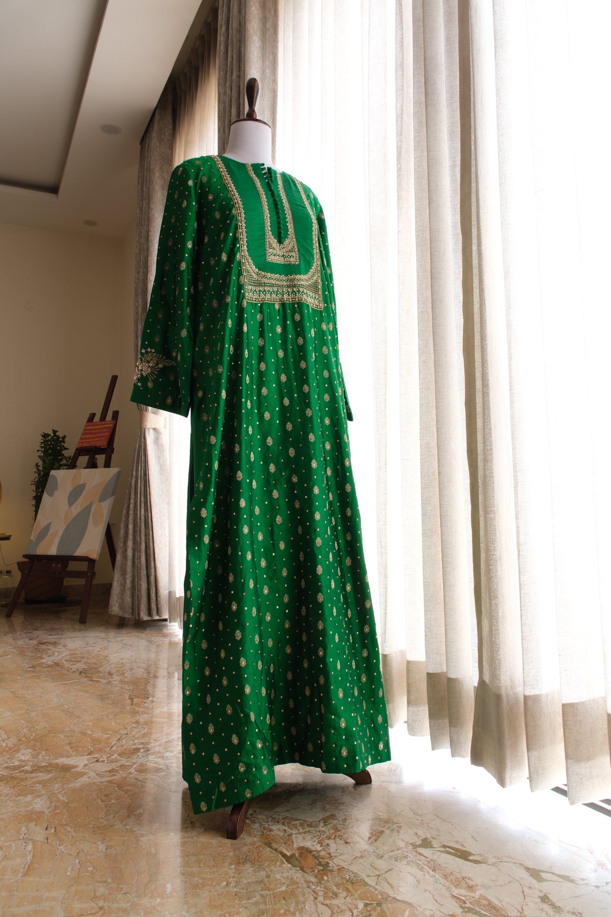 A long green kaftan dress with gold geometric patterns and a gold embroidered neckline, displayed on a mannequin in a bright room with white curtains.
